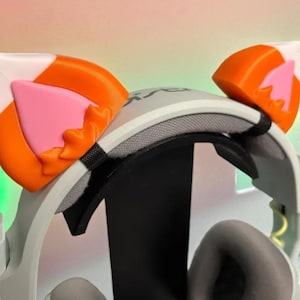 May include: A white and gray VR headset with orange and pink cat ear attachments. The headset has gray earcups and a black adjustable strap. The cat ears are orange with pink inner ears and a jagged edge.