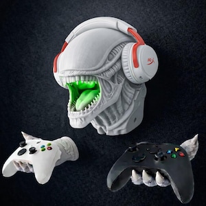 Könnte beinhalten: Eine graue Alien-Kopfskulptur mit einem leuchtend grünen Mund und Zunge, die weiße und rote Kopfhörer trägt. Darunter zwei Controller-Halter, einer weiß und einer schwarz, jeweils von einer geformten Hand gehalten.
