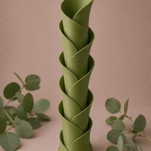 May include: A stack of five green, petal-shaped decorative vases. The vases are arranged in a spiral, with each petal overlapping the one below. The vases have a textured surface and are set against a neutral background with leafy sprigs.