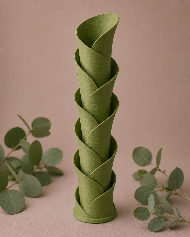 May include: A stack of five green, petal-shaped decorative vases. The vases are arranged in a spiral, with each petal overlapping the one below. The vases have a textured surface and are set against a neutral background with leafy sprigs.