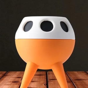 May include: A unique orange and white planter with three legs. The planter has a rounded orange base and a white top with three black circular openings. The planter is sitting on a wooden surface.