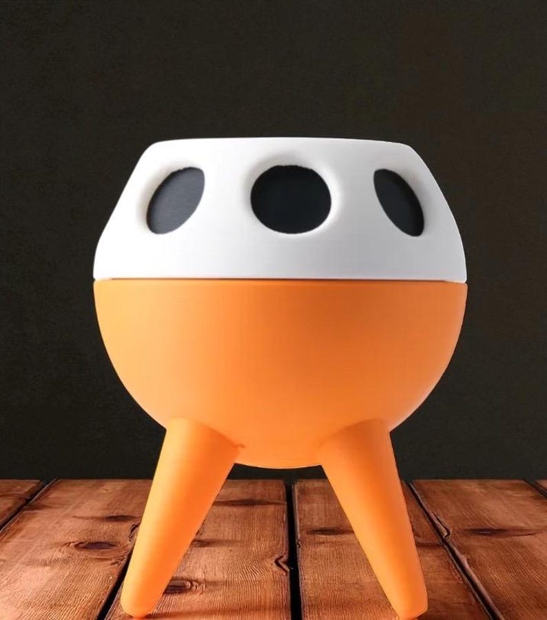 May include: A unique orange and white planter with three legs. The planter has a rounded orange base and a white top with three black circular openings. The planter is sitting on a wooden surface.