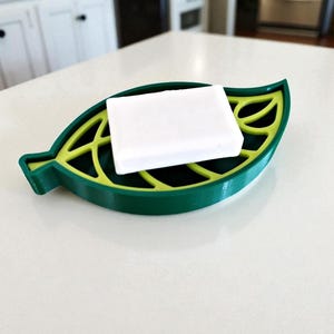 May include: A green and yellow leaf-shaped soap dish holding a white bar of soap. The dish has a detailed leaf design with a green outer edge and a yellow interior. The soap is rectangular and sits neatly inside the dish.
