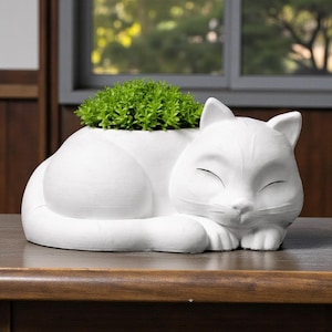 May include: A white ceramic cat-shaped planter, with a sleeping cat design. The planter holds a small, vibrant green plant. The cat is resting on a wooden table. The planter is a decorative item for home decor.