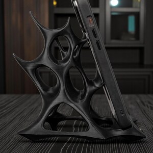 May include: Black phone stand with an intricate, spiky design. The stand holds a black smartphone, angled for viewing. The stand is made of a dark material and sits on a dark wood surface. The design is modern and artistic.