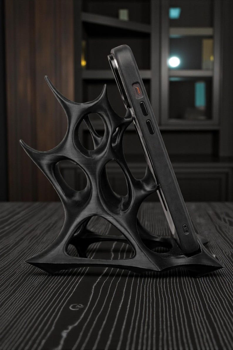 May include: Black phone stand with an intricate, spiky design. The stand holds a black smartphone, angled for viewing. The stand is made of a dark material and sits on a dark wood surface. The design is modern and artistic.