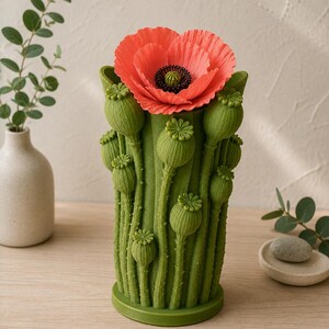 K&ouml;nnte beinhalten: Eine gr&uuml;ne, mohnf&ouml;rmige Vase mit einer leuchtend roten Mohnblume. Die Vase hat detaillierte gr&uuml;ne Mohnkapseln und -stiele. Die Blume hat eine schwarze Mitte und zarte Bl&uuml;tenbl&auml;tter. Die Vase ist ein dekoratives St&uuml;ck.