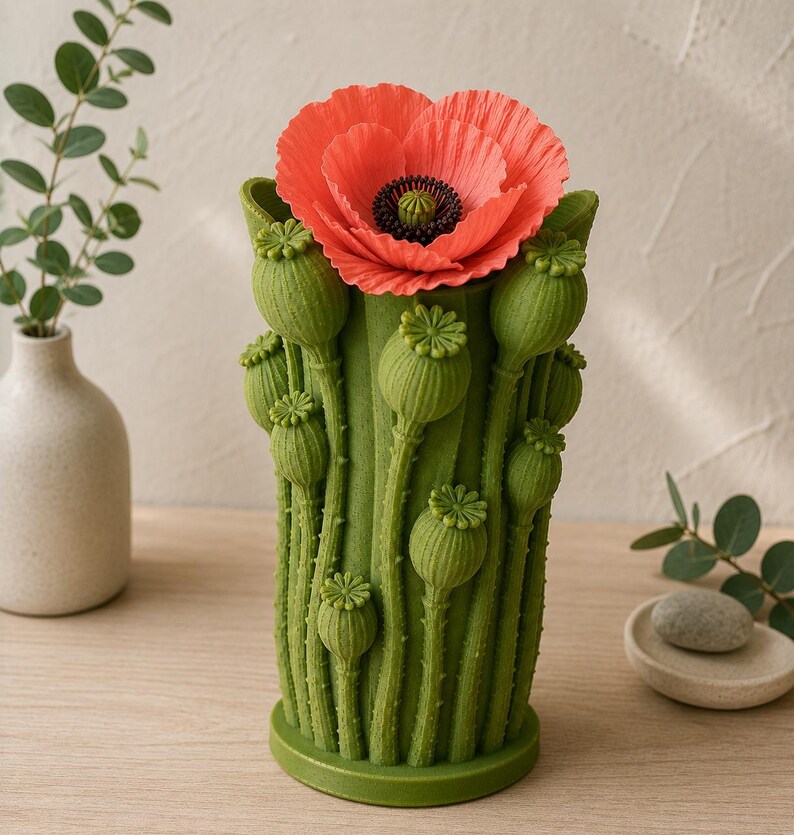 K&ouml;nnte beinhalten: Eine gr&uuml;ne, mohnf&ouml;rmige Vase mit einer leuchtend roten Mohnblume. Die Vase hat detaillierte gr&uuml;ne Mohnkapseln und -stiele. Die Blume hat eine schwarze Mitte und zarte Bl&uuml;tenbl&auml;tter. Die Vase ist ein dekoratives St&uuml;ck.