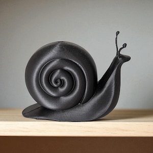 May include: A black, 3D-printed snail ornament. The snail features a spiraled shell and detailed antennae. The ornament is displayed on a light-coloured wooden surface against a neutral background. The snail is approximately 15 cm long.