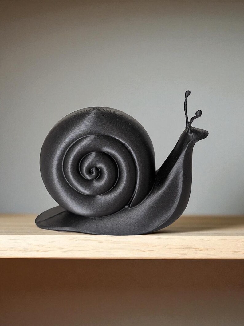 May include: A black, 3D-printed snail ornament. The snail features a spiraled shell and detailed antennae. The ornament is displayed on a light-coloured wooden surface against a neutral background. The snail is approximately 15 cm long.