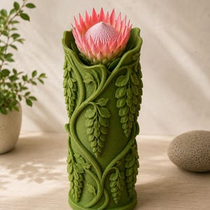 Puede incluir: Un jarr&oacute;n cil&iacute;ndrico verde con un dise&ntilde;o detallado de hojas, que sostiene una flor de protea rosa. El jarr&oacute;n tiene una superficie texturizada con enredaderas y hojas colgantes. La flor tiene una apariencia espinosa y en capas.