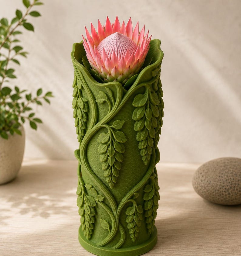 Puede incluir: Un jarr&oacute;n cil&iacute;ndrico verde con un dise&ntilde;o detallado de hojas, que sostiene una flor de protea rosa. El jarr&oacute;n tiene una superficie texturizada con enredaderas y hojas colgantes. La flor tiene una apariencia espinosa y en capas.