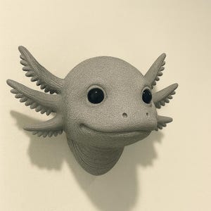 Puede incluir: Una escultura de ajolote gris con una expresi&oacute;n sonriente y ojos grandes y negros. La escultura tiene branquias detalladas y una superficie texturizada, montada en una pared de color neutro. El ajolote es una pieza &uacute;nica de decoraci&oacute;n del hogar.