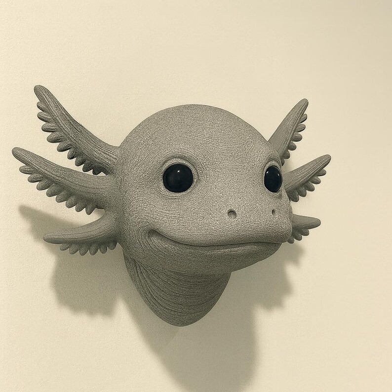 Puede incluir: Una escultura de ajolote gris con una expresi&oacute;n sonriente y ojos grandes y negros. La escultura tiene branquias detalladas y una superficie texturizada, montada en una pared de color neutro. El ajolote es una pieza &uacute;nica de decoraci&oacute;n del hogar.
