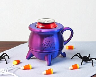 May include: A purple and blue cauldron-shaped drink holder with a skull and crossbones design. A red soda can sits inside the holder. Candy corn and black spiders are scattered around the cauldron on a white surface.