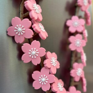 May include: A decorative garland of pink cherry blossom flowers with white centers. The flowers are attached to a brown, twig-like structure. The garland is displayed on a metallic surface.