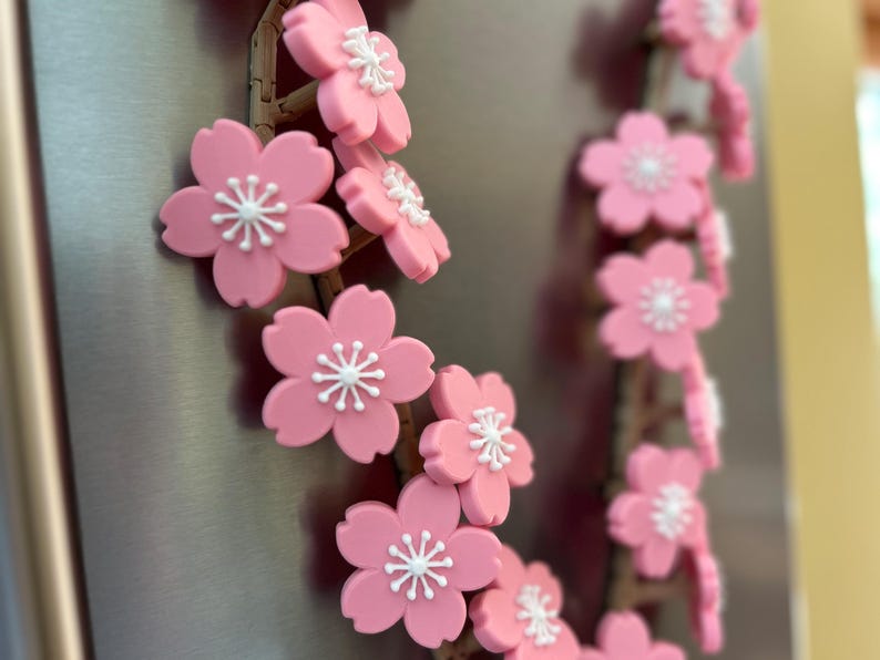 May include: A decorative garland of pink cherry blossom flowers with white centers. The flowers are attached to a brown, twig-like structure. The garland is displayed on a metallic surface.