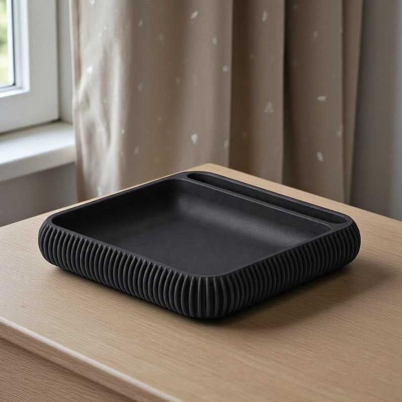 May include: A square, black tray with a ribbed, rounded edge. The tray has a small, rectangular compartment on one side. The tray is sitting on a light-colored wooden surface.