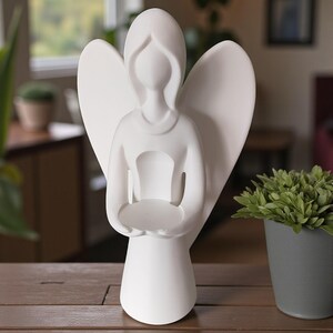 May include: A white ceramic angel figurine with outstretched wings and a circular platform held in its hands. The angel has a simple, elegant design and is displayed on a wooden surface. The figurine is a decorative item.