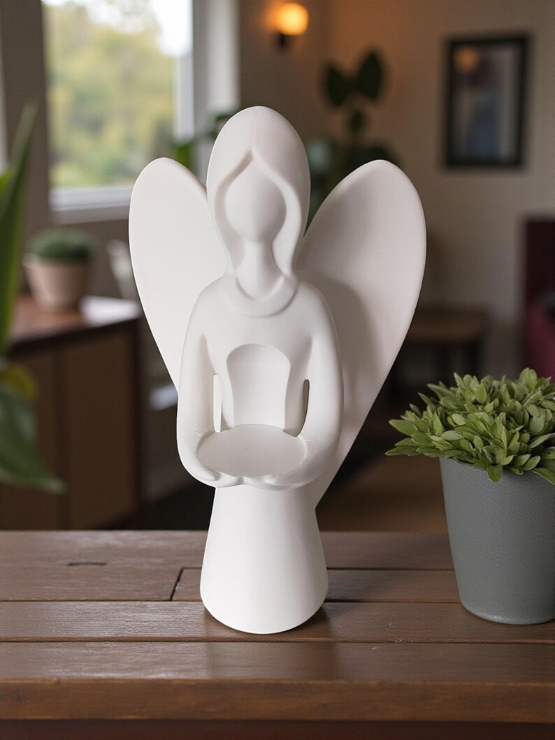 May include: A white ceramic angel figurine with outstretched wings and a circular platform held in its hands. The angel has a simple, elegant design and is displayed on a wooden surface. The figurine is a decorative item.