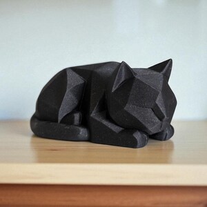 May include: A black geometric cat sculpture in a resting pose. The cat is made of angular shapes, giving it a modern, minimalist look. The sculpture is resting on a light-colored wooden surface.