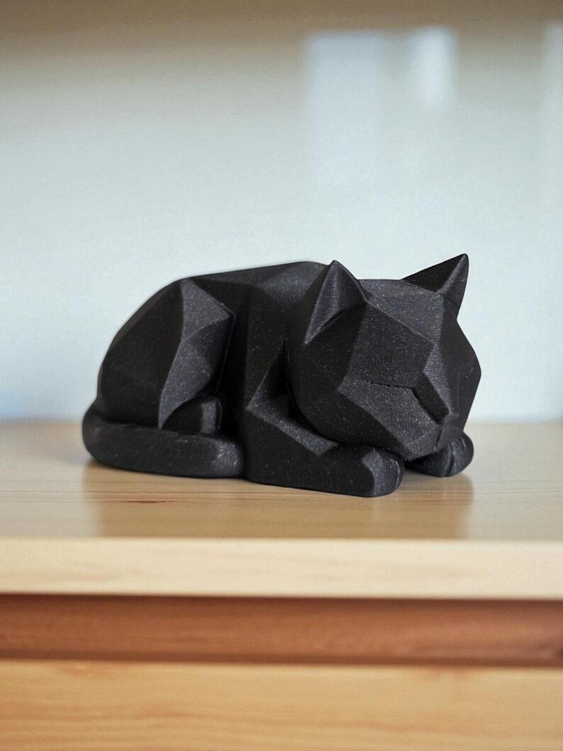 May include: A black geometric cat sculpture in a resting pose. The cat is made of angular shapes, giving it a modern, minimalist look. The sculpture is resting on a light-colored wooden surface.