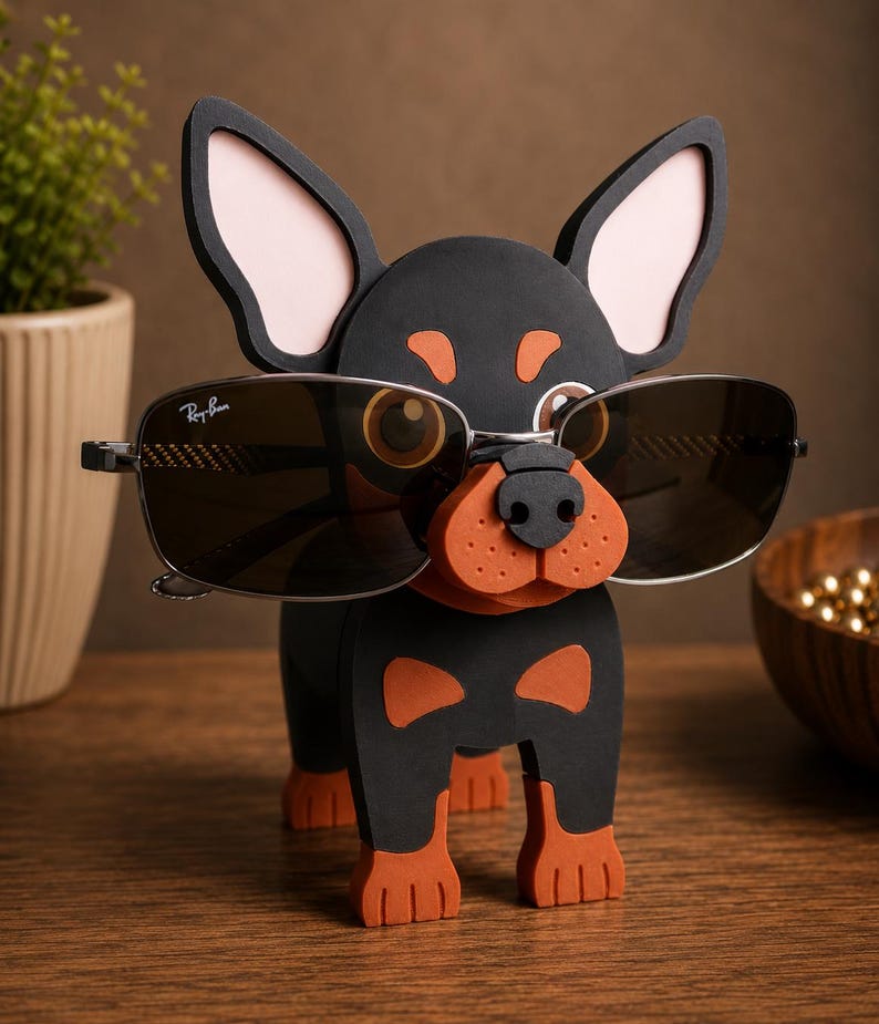 May include: A black and orange dog-shaped eyeglass holder with sunglasses. The holder has large ears with pink inner ears, and the sunglasses are held in place by the dog's nose. The sunglasses are Ray-Ban brand.