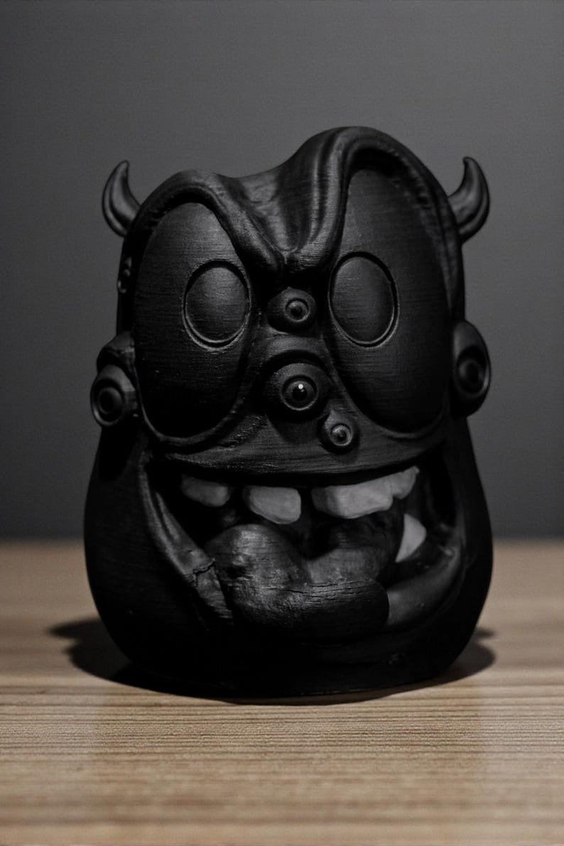 May include: A black, stylized sculpture of a monster with large eyes, horns, and a wide grin revealing white teeth. The sculpture has a textured surface and is set against a neutral background.