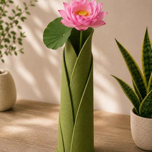 May include: A green felt vase shaped like leaves, holding a pink lotus flower with a green leaf. The vase is on a wooden surface, with plants in the background.