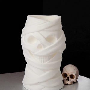 May include: A white, mummy-themed vase with a skull design, next to a small skull. The vase is wrapped in fabric-like material, creating a textured appearance. The background is black, and the objects are on a silver surface.