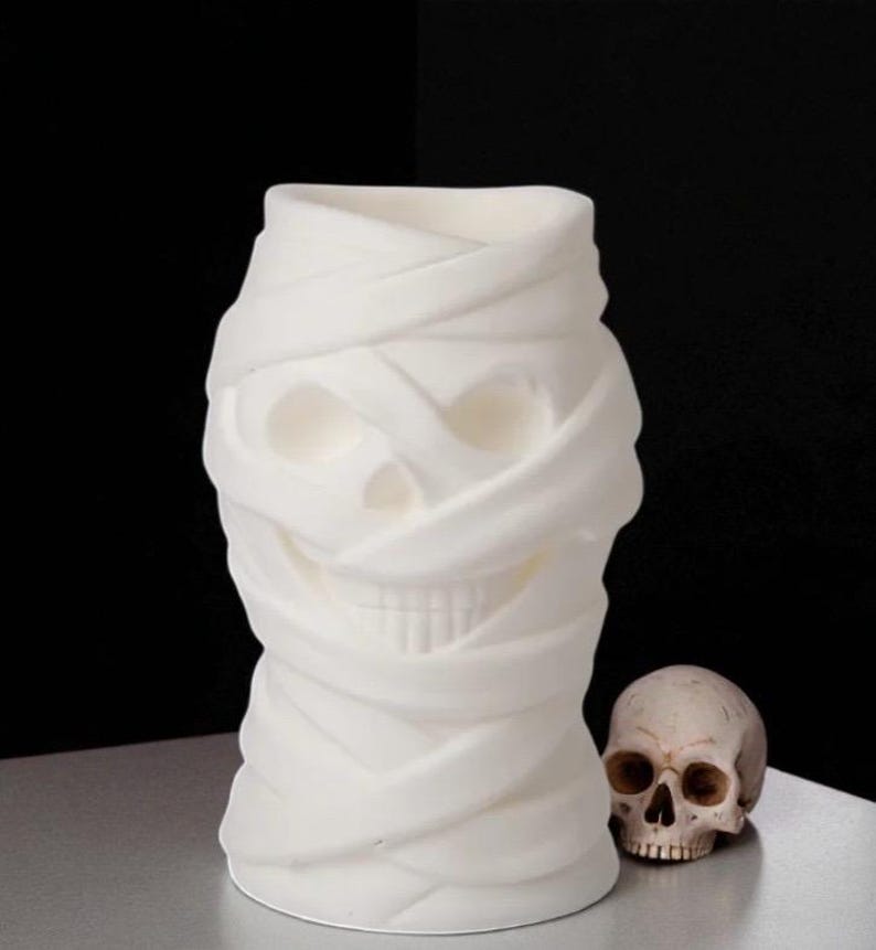 May include: A white, mummy-themed vase with a skull design, next to a small skull. The vase is wrapped in fabric-like material, creating a textured appearance. The background is black, and the objects are on a silver surface.