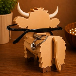 May include: A tan-colored wooden Highland cow-shaped eyeglasses holder with white horns. Black eyeglasses are resting on the cow's head. A set of keys hangs from the cow's body. The holder is on a wooden surface.