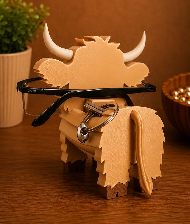 May include: A tan-colored wooden Highland cow-shaped eyeglasses holder with white horns. Black eyeglasses are resting on the cow's head. A set of keys hangs from the cow's body. The holder is on a wooden surface.