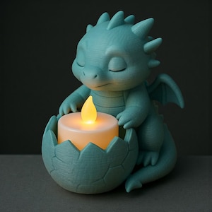 Puede incluir: Figura de drag&oacute;n turquesa sosteniendo una vela encendida en un soporte con forma de huevo. El drag&oacute;n tiene los ojos cerrados, peque&ntilde;as alas y una superficie texturizada. La vela emite una luz amarilla c&aacute;lida.