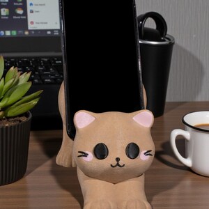 May include: A tan cat-shaped phone holder with pink inner ears and black eyes, holding a black smartphone. The holder sits on a wooden desk, with a laptop, a plant, and a coffee cup in the background.