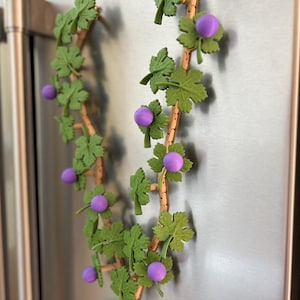 May include: A decorative grape vine garland with green leaves and purple grape clusters. The garland is attached to a stainless steel surface, likely a refrigerator door. The vine is made of a light brown material.