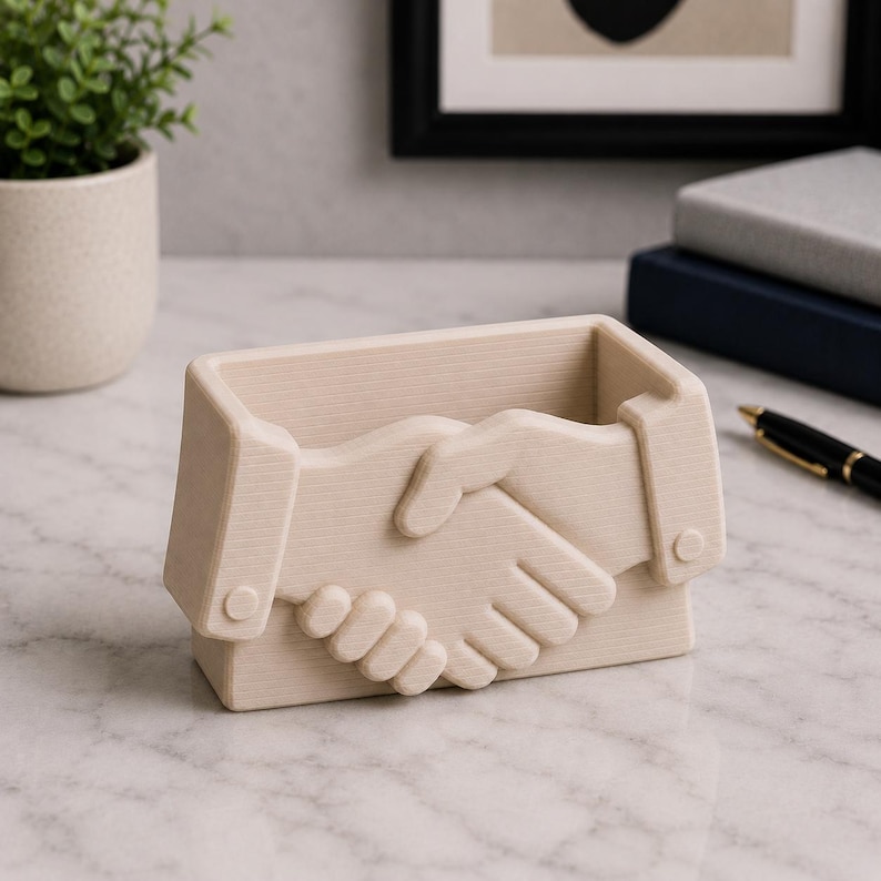 May include: A beige rectangular desk organizer with a handshake design. The organizer is made of a light-colored material and features a detailed carving of two hands clasped together. The design is on the front of the organizer.