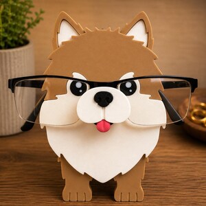 May include: A brown, white, and black dog-shaped eyeglasses holder with a pink tongue sticking out. The holder has black-framed glasses resting on it. The holder is made of a sturdy material and is designed to hold eyeglasses.