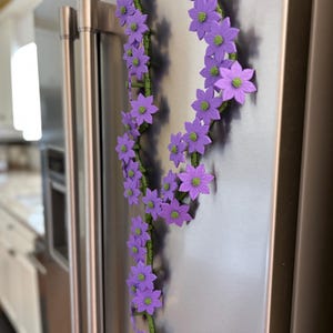 May include: A decorative floral garland with purple flowers and green stems is attached to a stainless steel refrigerator. The flowers have green centers. The garland is arranged in a vertical pattern.