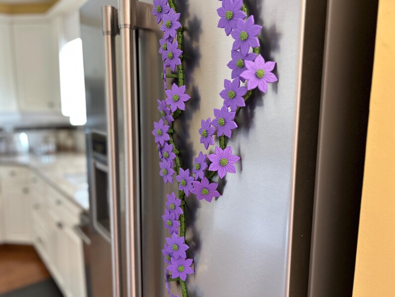 May include: A decorative floral garland with purple flowers and green stems is attached to a stainless steel refrigerator. The flowers have green centers. The garland is arranged in a vertical pattern.