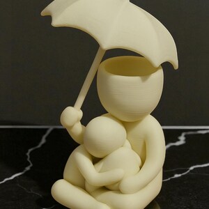 May include: A cream-colored figurine depicting a person holding an umbrella over another person. The figures are embracing. The umbrella is held by the larger figure, providing shelter. The sculpture is set against a dark, marbled surface.