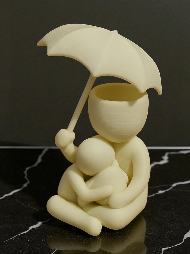 May include: A cream-colored figurine depicting a person holding an umbrella over another person. The figures are embracing. The umbrella is held by the larger figure, providing shelter. The sculpture is set against a dark, marbled surface.