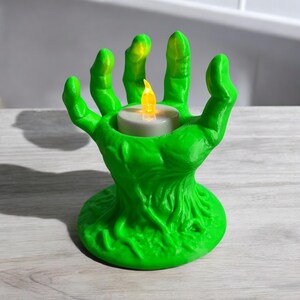 May include: A green, hand-shaped candle holder with a textured surface. The fingers cradle a white candle with a flickering yellow LED flame. The base has a textured, organic design, creating a spooky, Halloween-themed aesthetic.