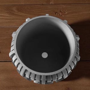 May include: A gray, cylindrical planter with a decorative, geometric design around the rim. The interior is dark gray, and a small white circle is visible at the bottom. The planter sits on a wooden surface.