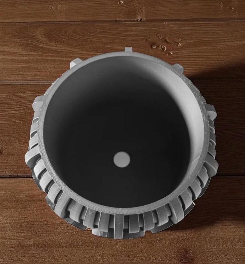 May include: A gray, cylindrical planter with a decorative, geometric design around the rim. The interior is dark gray, and a small white circle is visible at the bottom. The planter sits on a wooden surface.