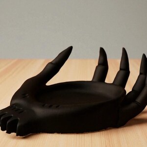 May include: A matte black hand-shaped decorative dish. The dish features a detailed design of a hand with long, pointed fingers and a shallow bowl-shaped palm. The dish is placed on a light-colored wooden surface.