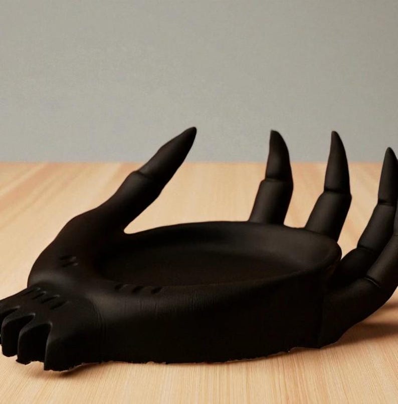 May include: A matte black hand-shaped decorative dish. The dish features a detailed design of a hand with long, pointed fingers and a shallow bowl-shaped palm. The dish is placed on a light-colored wooden surface.