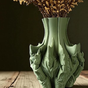 May include: A sage green decorative vase with a unique, textured design. The vase has a flared top and a detailed, organic pattern around the base. Dried flowers are arranged inside the vase, adding a natural element to the composition.
