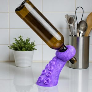 K&ouml;nnte beinhalten: Ein lila Tentakel-Weinflaschenhalter, der eine braune Glasflasche tr&auml;gt. Der Halter hat Saugnapfdetails und einen Ring zur Aufnahme der Flasche. Der Hintergrund umfasst eine wei&szlig; geflieste Wand, eine kleine Pflanze und K&uuml;chenutensilien.