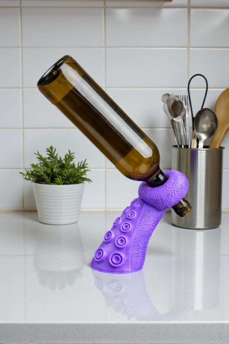 K&ouml;nnte beinhalten: Ein lila Tentakel-Weinflaschenhalter, der eine braune Glasflasche tr&auml;gt. Der Halter hat Saugnapfdetails und einen Ring zur Aufnahme der Flasche. Der Hintergrund umfasst eine wei&szlig; geflieste Wand, eine kleine Pflanze und K&uuml;chenutensilien.